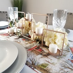 May include: A gold metal candle holder with five candle holders. The candle holder is on a table with a white tablecloth and a red and white patterned table runner. There are white candles in each of the candle holders.