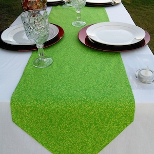 May include: A green table runner with a white leaf pattern. The runner is on a white tablecloth with red and white chargers and clear glass glasses.
