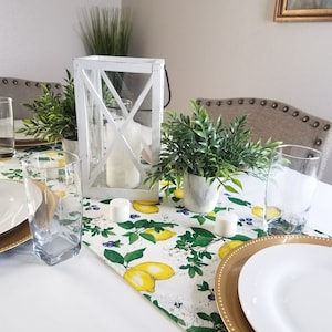 May include: A white table setting with a lemon-themed table runner, gold chargers, white plates, and clear glasses. There are two small potted plants and a white lantern with candles on the table.