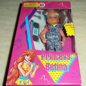 May include: A pink box with a white and blue doll inside. The doll has blonde hair and is wearing a pink shirt and blue overalls. The box has the words "Princess Betina" on it.
