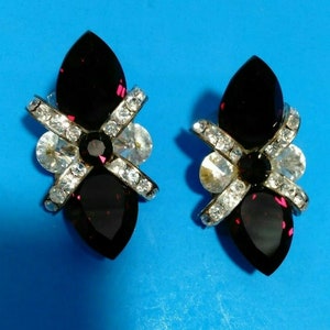 May include: A pair of clip-on earrings with a silver tone setting and three dark purple stones. The stones are set in a cross pattern and surrounded by clear rhinestones.