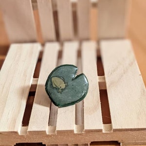 May include: A green and yellow frog sitting on a green lily pad. The lily pad is made of resin and has a glossy finish.