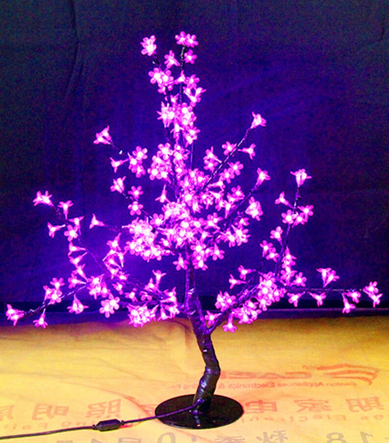 May include: A pink LED cherry blossom tree with a black base and a USB cord. The tree is lit up and the lights are on.