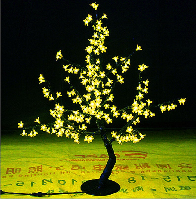 May include: A decorative artificial tree with a black trunk and branches covered in small yellow LED lights, resembling a cherry blossom tree.