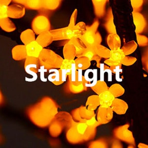 May include: A string of warm white fairy lights shaped like flowers. The lights are strung on a black wire and the text "Starlight" is visible in the center of the image.