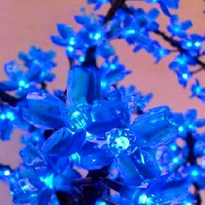 May include: A close-up of a blue artificial flower branch with small, delicate blossoms. The flowers are made of plastic and have a shiny, iridescent finish. The branch is perfect for adding a touch of colour and whimsy to any room.