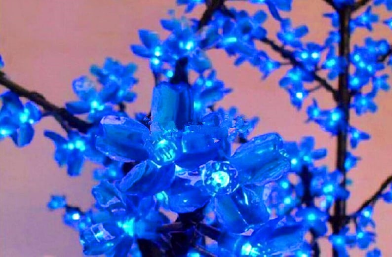 May include: A close-up of a blue artificial flower branch with small, delicate blossoms. The flowers are made of plastic and have a shiny, iridescent finish. The branch is perfect for adding a touch of colour and whimsy to any room.