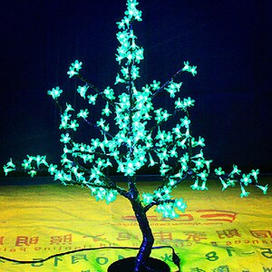 May include: A decorative LED light-up tree with a black base and green lights shaped like flowers. The tree is on a yellow surface with Chinese characters.