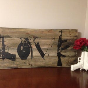 May include: A wooden sign with the word "LOVE" spelled out in black paint. The letters are formed by images of guns, a grenade, and a knife. The sign is mounted on a wall with red roses in a white vase shaped like a gun.