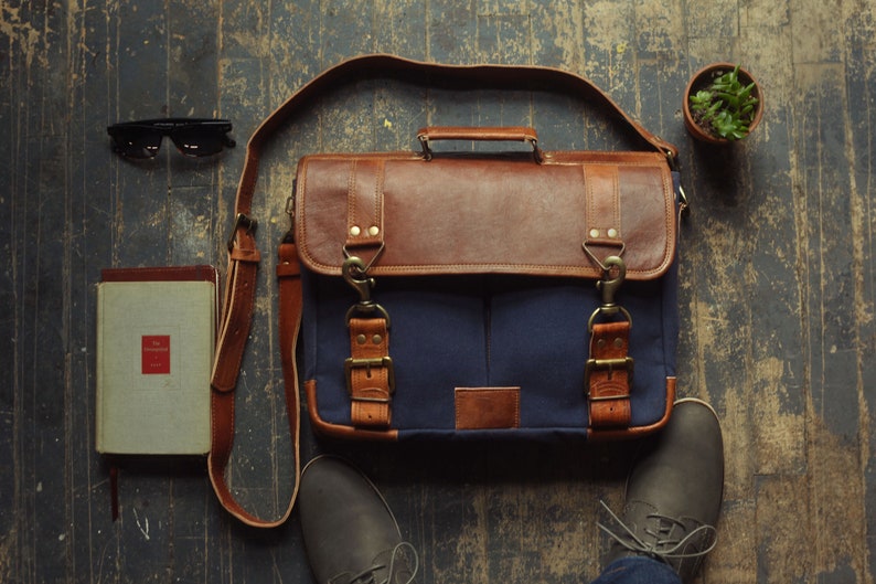 K&ouml;nnte beinhalten: Eine braune Ledertasche mit einem blauen Canvas-Klappen und Messing-Schnallen. Die Tasche hat einen langen verstellbaren Riemen und steht auf einem Holzboden mit einem Buch, einer Sonnenbrille und einer Topfpflanze.