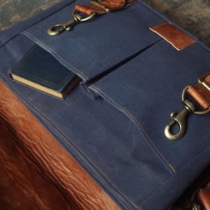 K&ouml;nnte beinhalten: Eine blaue Canvas-Messenger-Tasche mit braunen Lederakzenten und einer gro&szlig;en Fronttasche. Die Tasche ist offen und ein kleines Buch ist in der Tasche sichtbar.