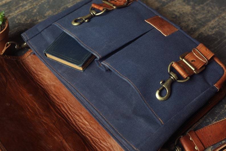 K&ouml;nnte beinhalten: Eine blaue Canvas-Messenger-Tasche mit braunen Lederakzenten und einer gro&szlig;en Fronttasche. Die Tasche ist offen und ein kleines Buch ist in der Tasche sichtbar.