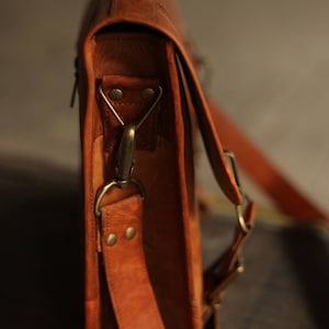 May include: A brown leather briefcase with a long strap and a brass buckle. The bag has a flap closure and a textured leather finish.