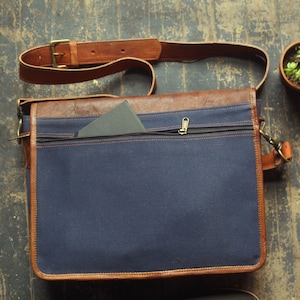 K&ouml;nnte beinhalten: Eine blaue Canvas- und braune Leder-Messenger-Tasche mit einer Rei&szlig;verschlusstasche und einem Klappverschluss. Die Tasche hat einen braunen Lederriemen.