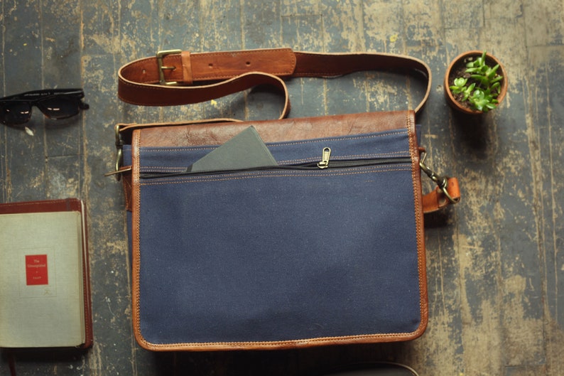 K&ouml;nnte beinhalten: Eine blaue Canvas- und braune Leder-Messenger-Tasche mit einer Rei&szlig;verschlusstasche und einem Klappverschluss. Die Tasche hat einen braunen Lederriemen.
