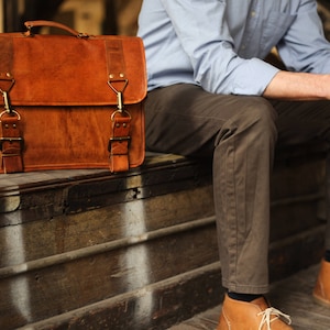 May include: A brown leather briefcase with brass buckles and a strap. The briefcase is sitting on a wooden surface.