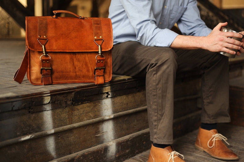 May include: A brown leather briefcase with brass buckles and a strap. The briefcase is sitting on a wooden surface.