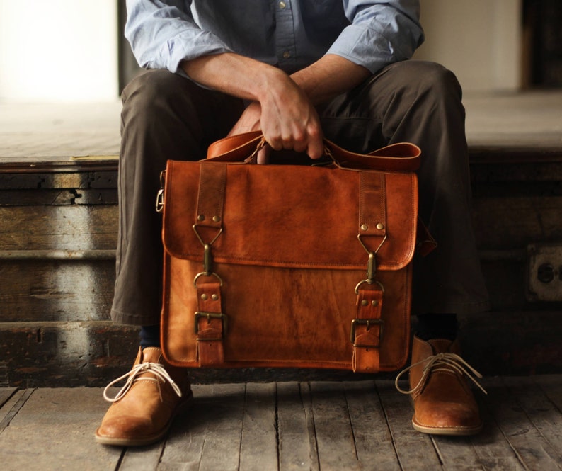 May include: A brown leather messenger bag with two adjustable straps and brass buckles. The bag has a flap closure and a top handle.