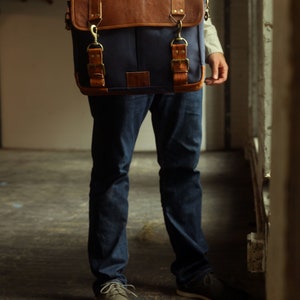 K&ouml;nnte beinhalten: Eine Person in blauen Jeans und braunen Lederschuhen tr&auml;gt eine blaue Canvas-Messenger-Tasche mit braunen Lederakzenten und Messingverschl&uuml;ssen.
