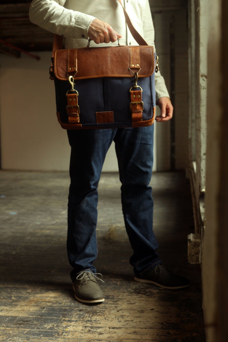 K&ouml;nnte beinhalten: Eine Person in blauen Jeans und braunen Lederschuhen tr&auml;gt eine blaue Canvas-Messenger-Tasche mit braunen Lederakzenten und Messingverschl&uuml;ssen.