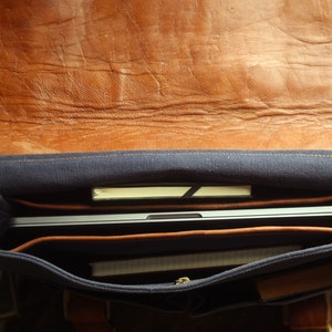K&ouml;nnte beinhalten: Eine braune Ledertasche mit blauer Canvas-Futter. Die Tasche ist offen und enth&auml;lt einen Laptop, ein Notizbuch und andere Gegenst&auml;nde.