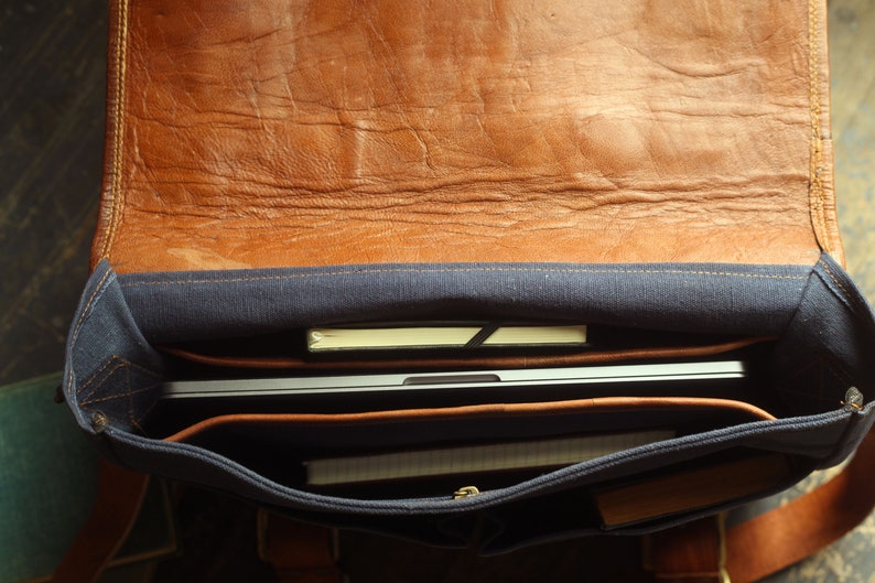 K&ouml;nnte beinhalten: Eine braune Ledertasche mit blauer Canvas-Futter. Die Tasche ist offen und enth&auml;lt einen Laptop, ein Notizbuch und andere Gegenst&auml;nde.