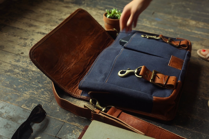K&ouml;nnte beinhalten: Eine blaue Canvas-Messenger-Tasche mit braunen Lederakzenten und Riemen. Die Tasche ist offen und das Innere ist mit braunem Leder ausgekleidet.
