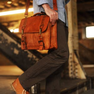 May include: A brown leather messenger bag with a long adjustable strap. The bag has two buckles and a flap closure. The bag is worn by a person wearing brown pants and a light blue shirt.