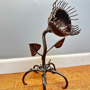 Op de afbeelding: Een metalen sculptuur van een vleesetende plant met een grote open mond gemaakt van schroeven. De plant heeft een lange stengel en twee bladeren. De basis van de plant is gemaakt van gedraaid metaal en heeft zes poten.