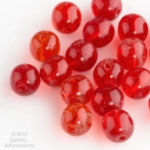 May include: A group of red glass beads with a speckled texture. The beads are round and have a hole in the center.