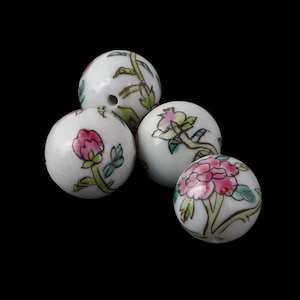 May include: Four white ceramic beads with floral designs. Each bead features hand-painted pink flowers, green leaves, and stems. The beads are round with a hole for stringing, set against a black background.
