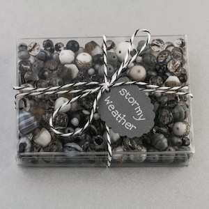 Puede incluir: Una caja de plástico transparente llena de cuentas de vidrio negras, blancas y grises. La caja está atada con un cordel y una etiqueta negra que dice "tiempo tormentoso".