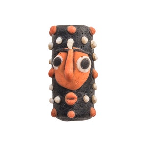 May include: A cylindrical ceramic bead with a face design. The bead is black with orange and white accents. The face has large eyes, a prominent nose, and a small mouth. The bead is decorated with small, raised dots. A unique craft item.