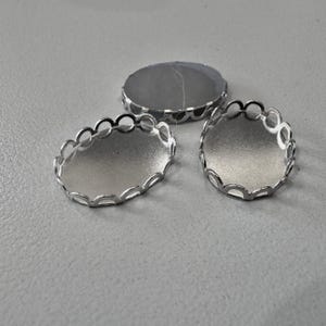 May include: Three silver-colored oval bezel trays for jewelry making. The trays have a scalloped edge design and a flat, reflective surface. The trays are empty and ready for use in crafting projects.