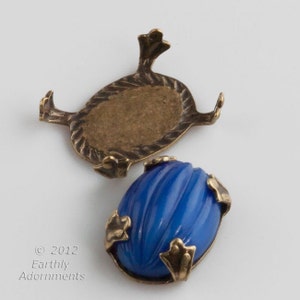 Puede incluir: Un cabujón de vidrio azul con textura acanalada, engastado en un engaste de latón con cuatro patas decorativas. El engaste se muestra por separado.