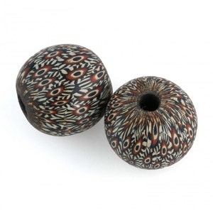 Puede incluir: Dos cuentas redondas de vidrio negro y blanco con un patrón geométrico marrón y rojo. Las cuentas tienen un agujero en el centro.