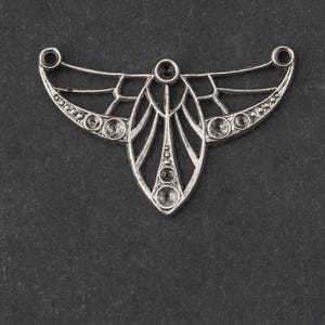 Puede incluir: Un colgante de metal plateado con un diseño Art Déco. El colgante presenta una forma central de lágrima con líneas radiales y acentos circulares. Pequeñas piedras transparentes están engastadas en los bordes y en el centro.