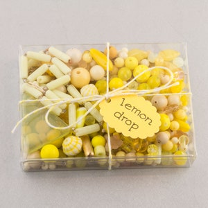 Puede incluir: Una caja de plástico transparente llena de cuentas amarillas y blancas. La caja está atada con un cordel y una etiqueta amarilla que dice "lemon drop".
