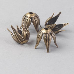 Könnte beinhalten: Vier antike, messingfarbene Metallperlenkappen, jede in Form einer stilisierten Blume mit detaillierten blattartigen Blütenblättern. Die Kappen haben oben ein kleines Loch und sind für Schmuckherstellung oder Bastelprojekte gedacht.