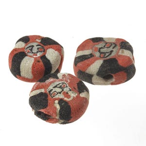 May include: Three round ceramic beads with a hole through the centre. The beads are decorated with black, white and red stripes and a stylised face in the centre.