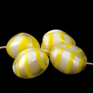 May include: Four glass beads with yellow and white stripes. The beads are oval shaped and have a glossy finish.