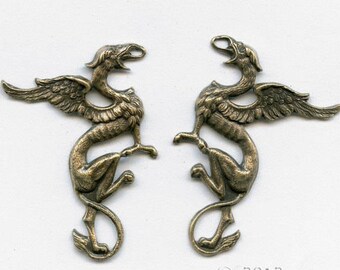 Op de afbeelding: Twee antieke goudkleurige metalen griffin-bedels met uitgespreide vleugels. De griffioenen kijken elkaar aan.