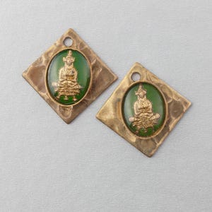 Puede incluir: Dos colgantes de color latón con forma de diamante y textura martillada. Cada colgante presenta un óvalo verde con una figura dorada de Buda sentado. Los colgantes tienen un pequeño agujero en la parte superior para colgar.