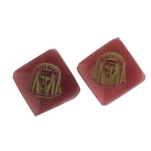 May include: Two square-shaped, pink glass tiles with a green, raised image of a face with a headdress.