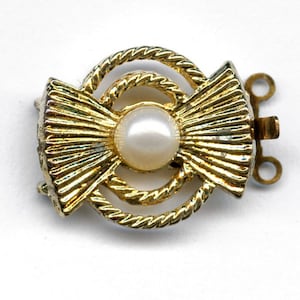 Puede incluir: Cierre de metal dorado con una perla falsa en el centro y un diseño de cuerda. El cierre tiene dos bucles para sujetar un collar o una pulsera.
