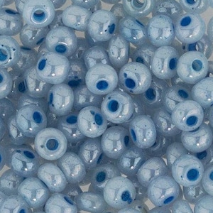Può includere: Primo piano di un mucchio di perline di vetro azzurro. Ogni perlina ha un foro blu scuro e una finitura lucida. Le perline sono rotonde, di forma leggermente irregolare, e sono adatte per la creazione di gioielli.