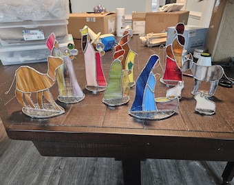 May include: A collection of stained glass nativity figures featuring a camel, various robed characters in shades of blue, red, yellow, green, and pink, and a donkey. Each piece is constructed with soldered metal edges and stands on a base.
