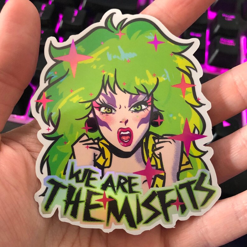 May include: A holographic sticker featuring a green-haired woman with pink and purple makeup, wearing a yellow and black outfit. The sticker has the text "We Are The Misfits" in a black and white font.