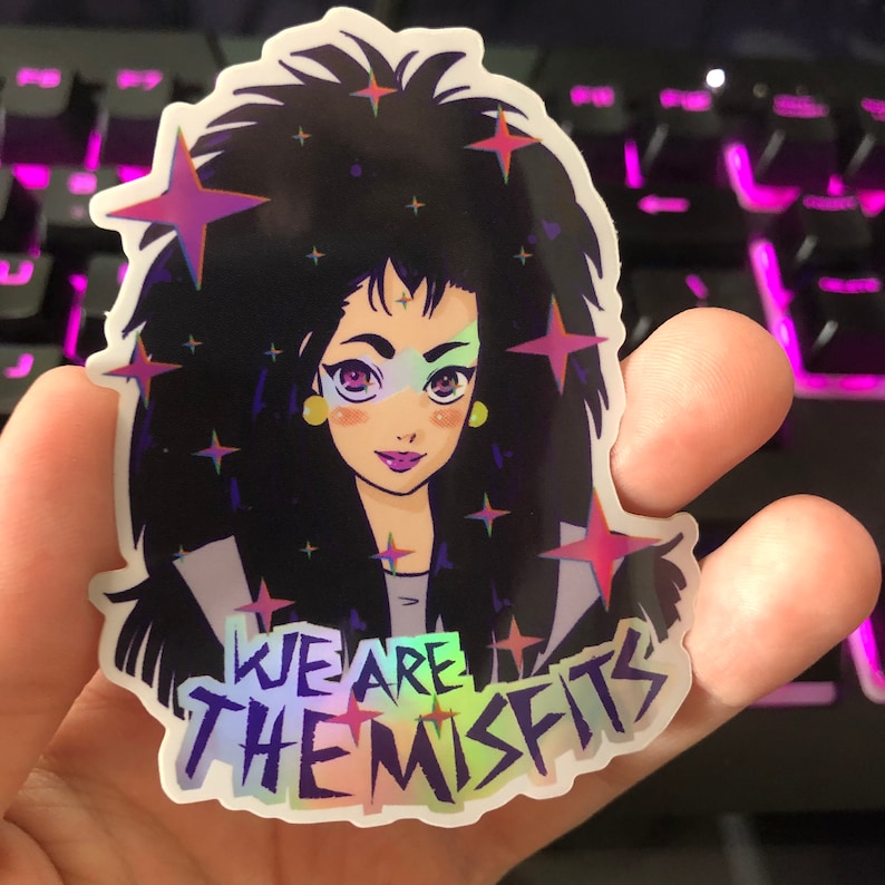 May include: A holographic sticker featuring a cartoon character with black hair, a pink and blue face, and yellow earrings. The character is surrounded by stars and the text "WE ARE THE MISFITS" is printed below.