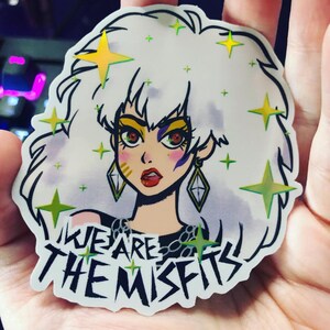 May include: A white sticker with a cartoon illustration of a woman with white hair and a black outfit. The woman has yellow and green stars around her head. The sticker has the text "We Are The Misfits" in black.